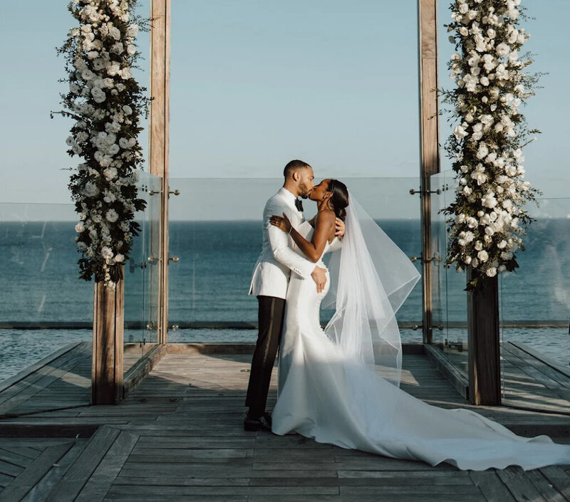 A bride and groom share a kiss at an oceanfront wedding ceremony. The groom is in a white tuxedo jacket, and the bride is wearing a form-fitting white gown with a long veil. Two large floral arrangements frame the couple, and the ocean is visible in the background. Keywords: luxury destination weddings, destination wedding planning, oceanfront wedding ceremony.