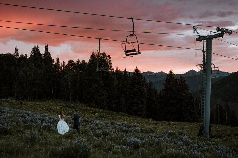 Wedding couple walking hand in hand through a field of wildflowers, with a ski lift and sunset drenched mountain background near Crested Butte, Colorado.