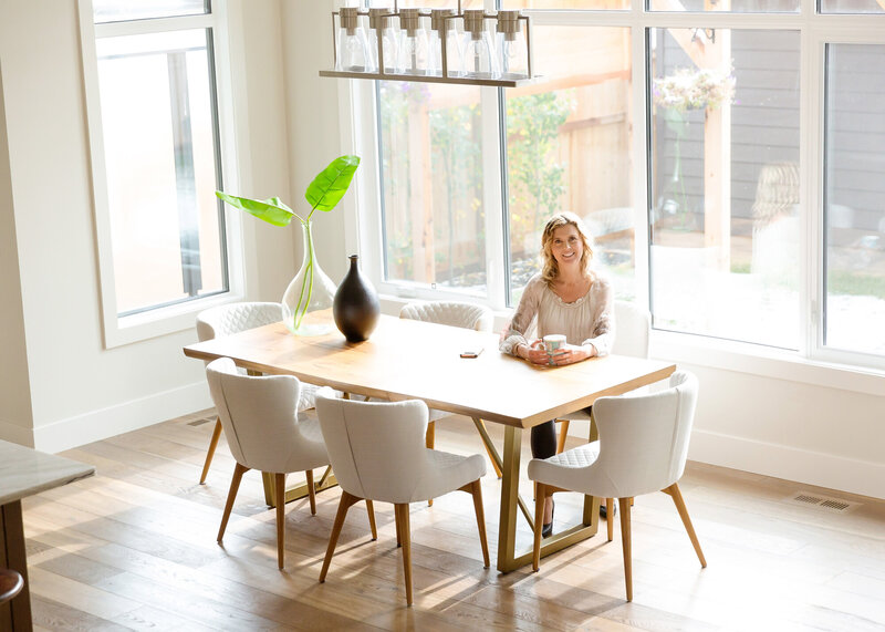 Calgary brand storytelling photoshoot of a woman sitting at a modern wooden dining table in a bright, airy home. She holds a coffee mug and smiles warmly, surrounded by elegant white chairs and minimalist decor. Large windows let in natural light, showcasing an inviting space with a view of the backyard garden.