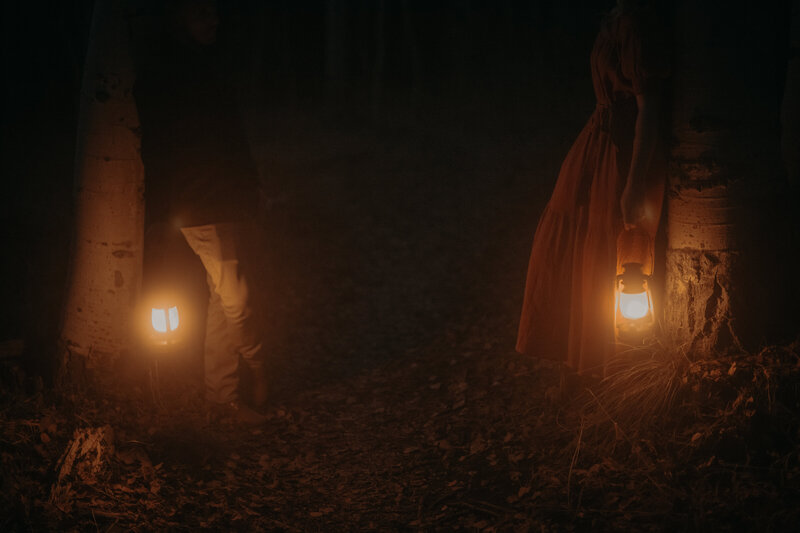 Two people standing against two trees face each other holding lanterns at their sides. It is dark and the lanterns are the most visible thing.