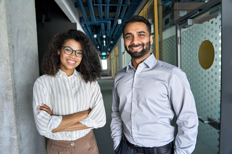 An Arab man and a Black woman are colleagues. They are wearing business casual and stand in a hallway smiling. 