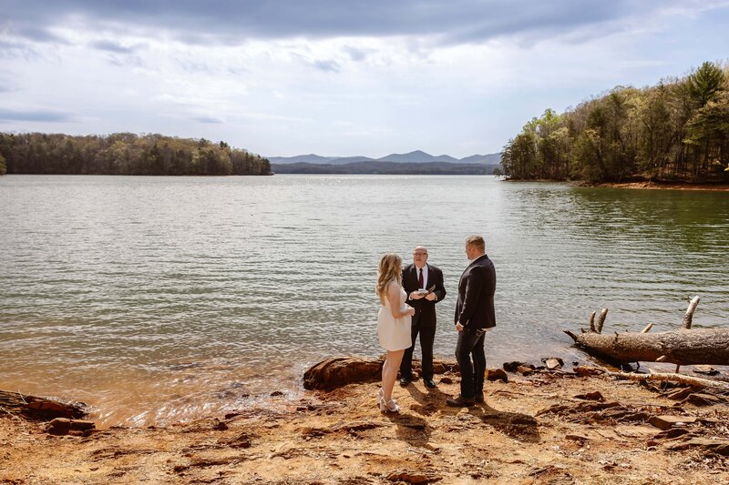 A bride and groom are walking on rocks at the base of a large tumbling waterfall. The bride is in a flowy white dress with a long veil and the groom is wearing tan pants with a red button up shirt and an orange leather jacket. The groom is holding the brides hand helping guide her along the rocks.
