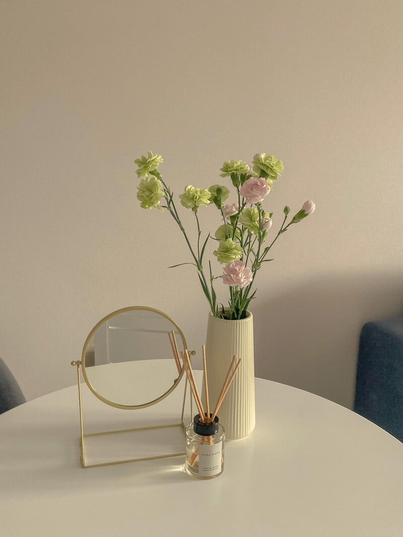 Green and pink flowers in a modern vase sit on desktop aside a modern gold mirror and room fragrance.