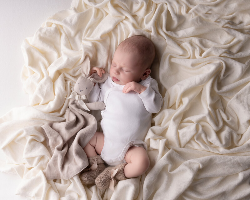 Baby i hvit body ligger på kremfarget tekstil med koseklut under fotografering i studio nær Oslo.
