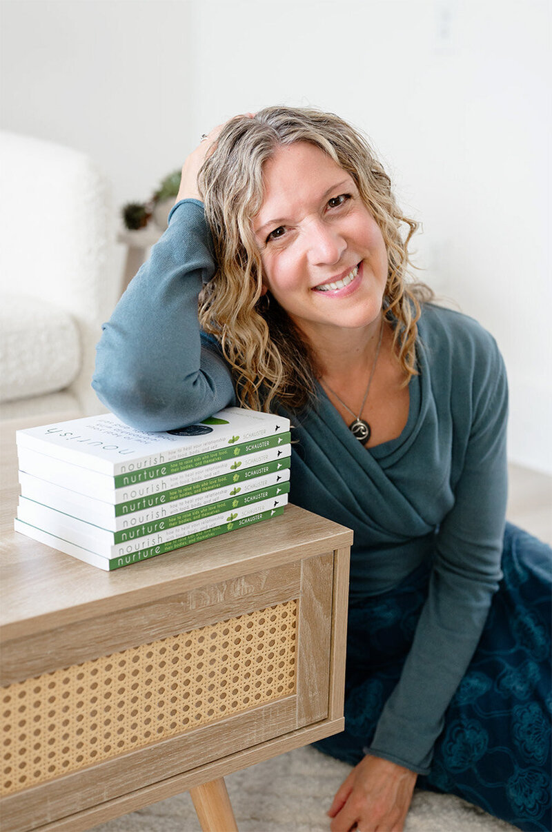 Heidi Schauster with curly hair leans on a stack of books titled "Nourish." She wears a teal top, sitting by a wooden table in a cozy, light room.
