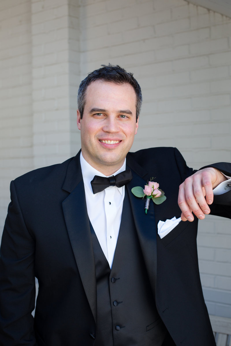 Groom in black tuxedo stands with arm bent with pink peony boutonniere