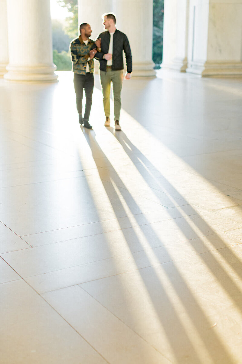 A gay couple walks arm in arm through the Jefferson Memorial in Washington, D.C., bathed in golden sunlight. Their long shadows stretch across the marble floor, emphasizing the warmth and intimacy of the moment. The soft focus captures their laughter and connection, framed by the monument's grand columns and architecture.