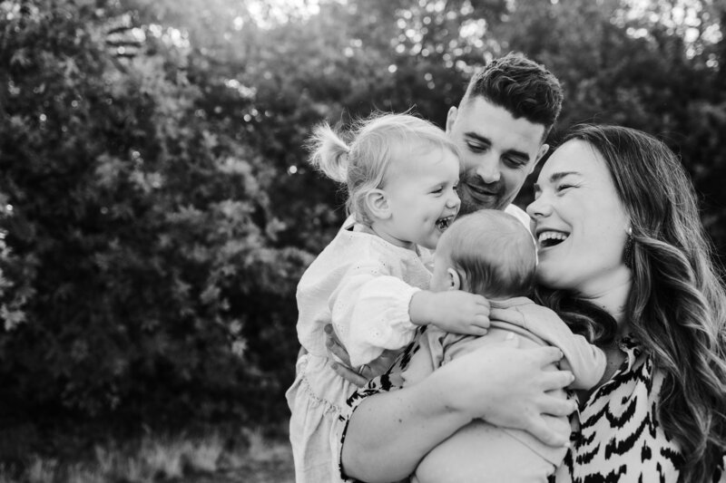 Gezin geeft elkaar een knuffel waarbij dochter hard lacht en papa en mama trots toekijken zwartwit