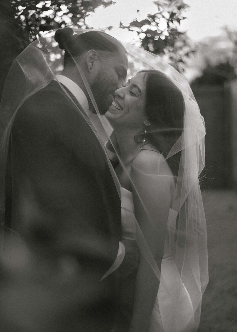 Groom whispering in brides ear with veil over them and laughing