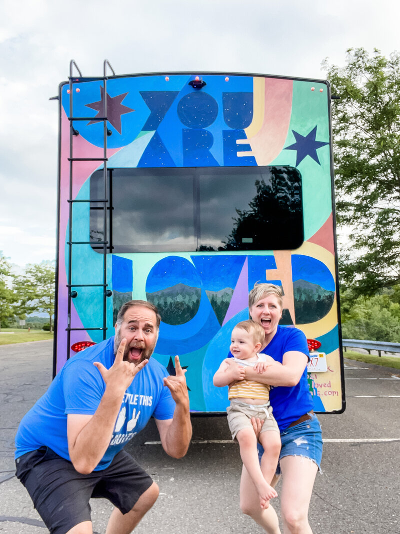 Mark and Kristin Palkoner posed with son in front of their 5th Wheel RV with You Are Loved mural by Alex Cook on back.
