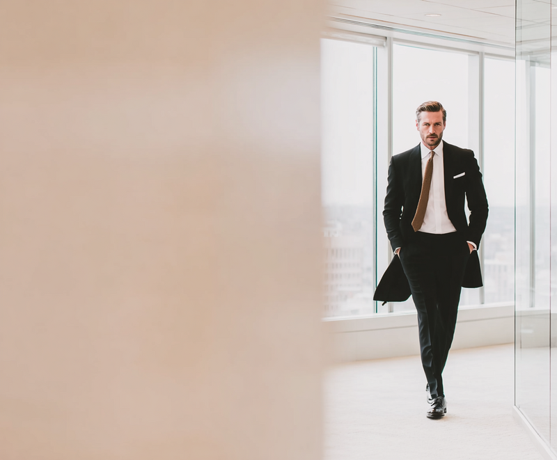 Stylish man in a tailored black suit walking through a modern glass corridor, symbolizing confidence, clarity, and the personal reinvention journey offered in Jasmina Kimova’s Genius Code Masterclass for visionary men and aligned creators.