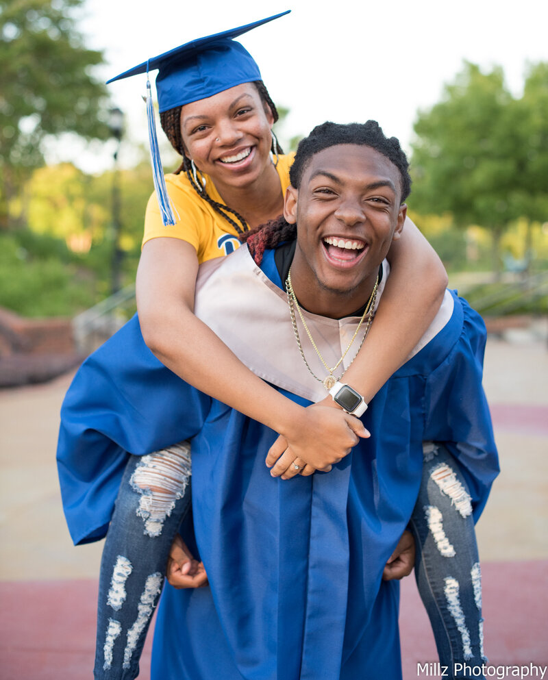 a high school senior dressed in his graduation gown carrying a woman wearing a graduation cap on his back.. photographed by Millz Photography in Greenville, SC