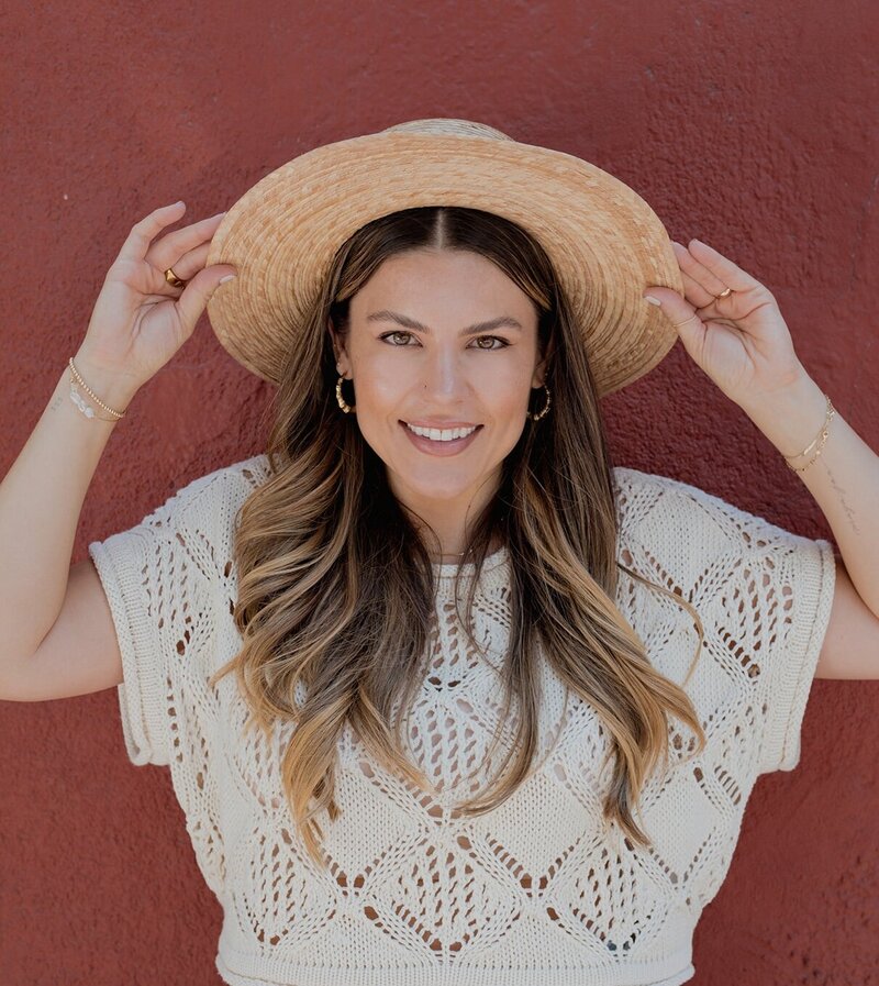 Natalie Bernacchi smiling in front of a red wall, holding a straw hat and wearing a cream knit top—radiating grounded joy