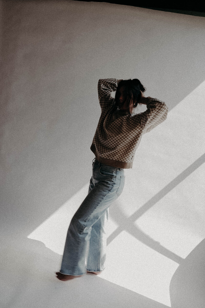 Tia Lorae wearing checkered brown sweater and blue jeans fixing hair in front of white backdrop