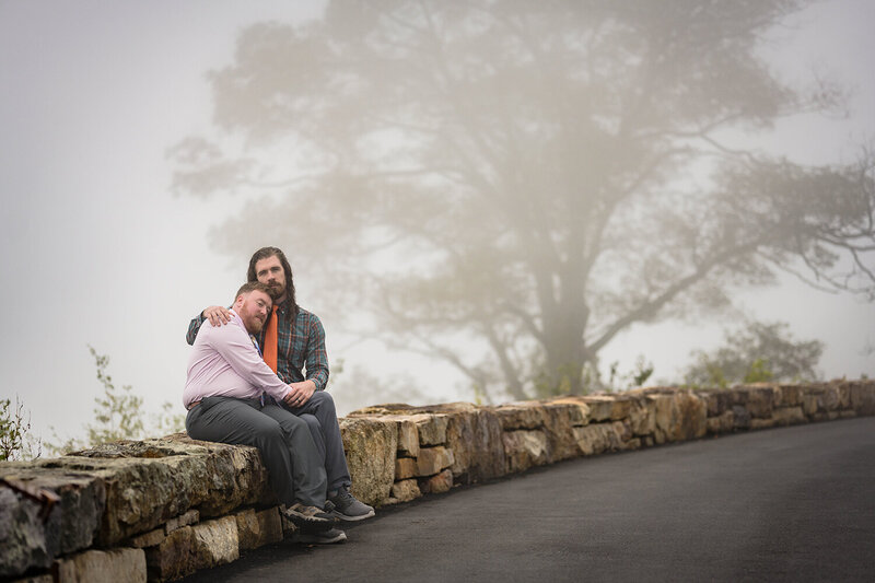An LGBTQ+ couple sit on a stone barrier at Loft Mountain Overlook in Shenandoah National Park. The background shows a large tree slightly covered by a thick cloud of fog.