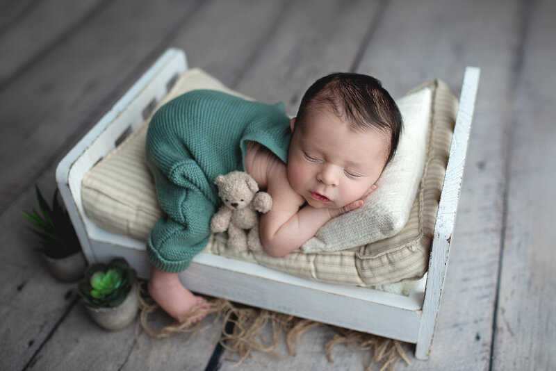 Newborn baby boy sleeping on a small bed wearing green overalls and holding a bear | CT Newborn Photographer Elizabeth Frederick Photography