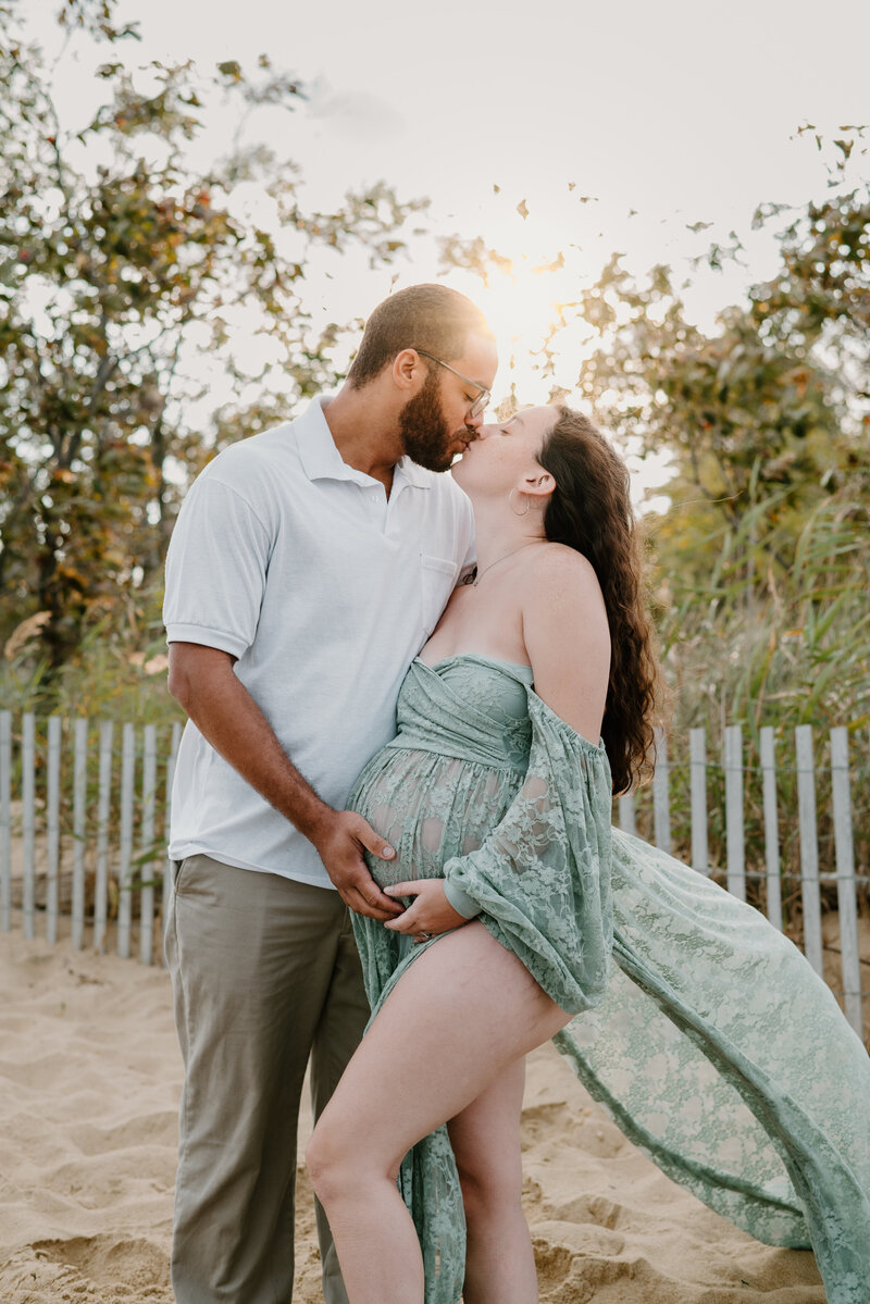 Pregnant mom in green lace dress holding her belly while kissing husband who is wearing white and tan while holding belly on a beach in Stevensville Maryland  photographed by Bethany Simms Photography