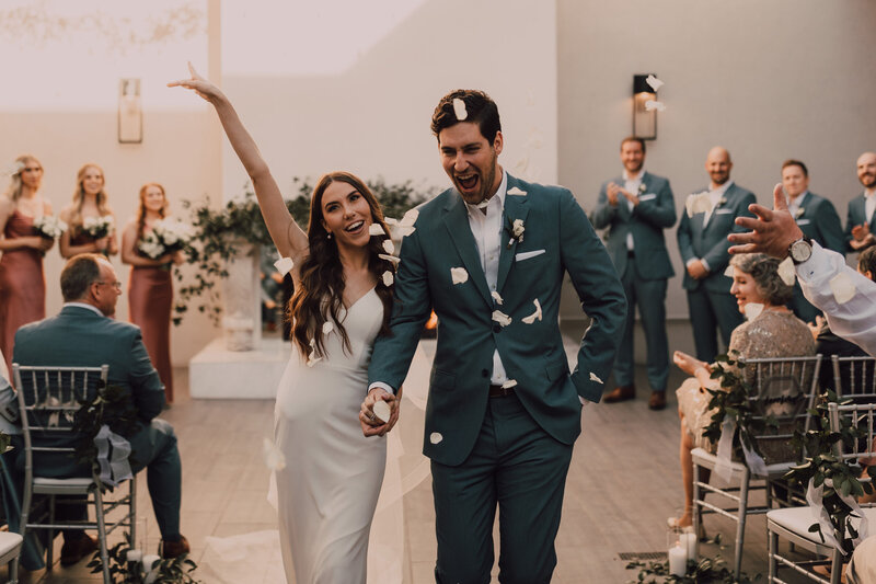 A bride and groom celebrate as they walk back down the aisle surrounded by falling flower petals at SoHo63, a Chandler wedding and event venue. The groom beams while the bride raises her arm in excitement, capturing the joyful energy of a just-married moment in SoHo63’s elegant ceremony space.