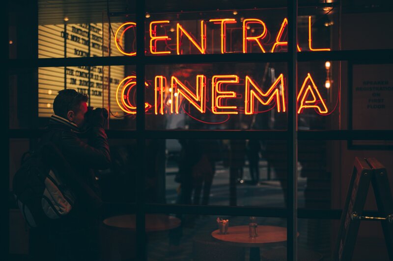 A person with a backpack takes a photograph of a neon sign that reads 'CENTRAL CINEMA' in vibrant red-orange letters, displayed in a window. The scene is set in a dimly lit environment, enhancing the glow of the neon, with the reflection of bustling street life visible in the glass.