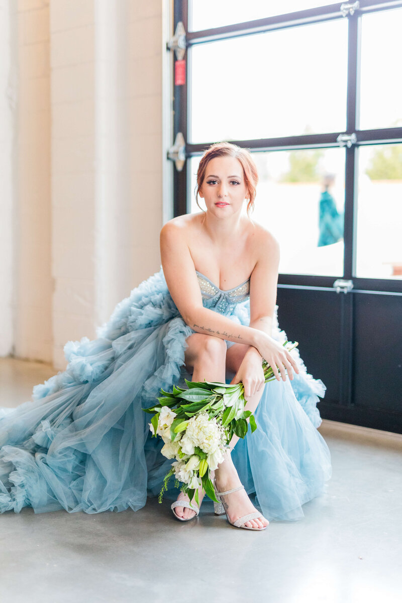 A bride in a dusty blue dress sits on a stool with her legs crossed looking at the camera. She is holding a bunch of roses in one hand as it's draped over her legs.