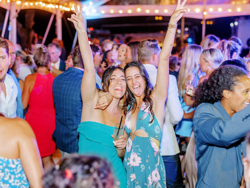 Two wedding guests posing with peace signs and their tongues out whilst on the dance floor with guests dancing around them at a New England tented wedding
