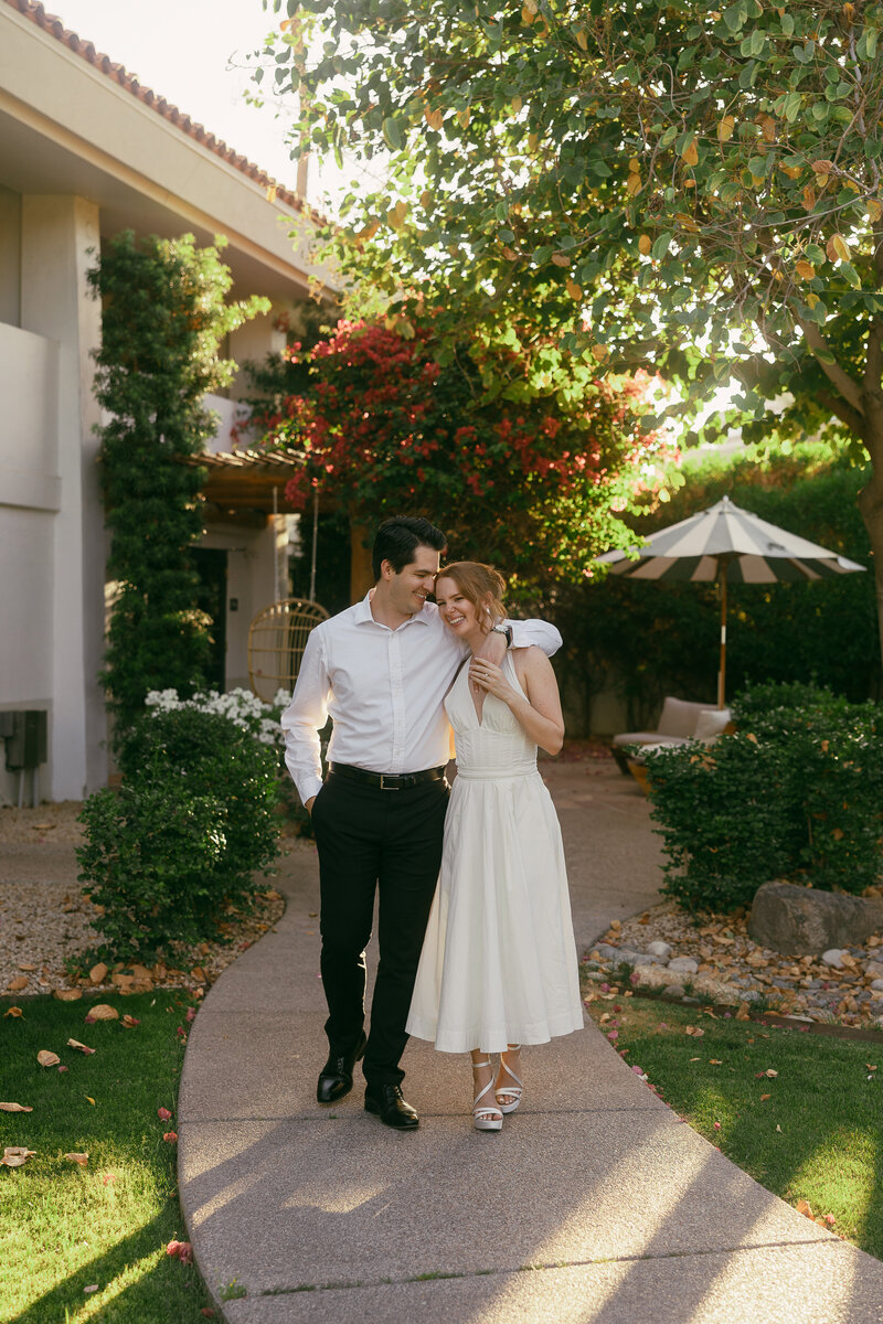 A couple with their arms around each other walk on a sidewalk surrounded by greenery and sunlit plants.