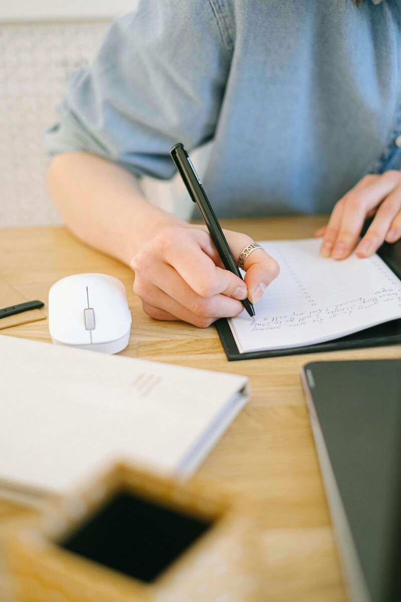 Image of an upper body of a woman writing in a notebook next to a computer mouse