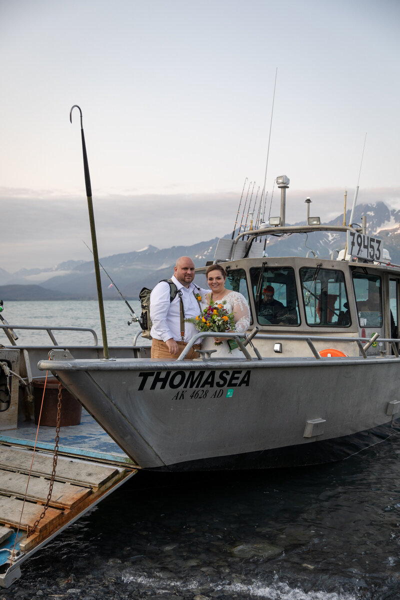 Bride and groom stand on a fishing boat named 'Thomasea,' docked in a glacial bay with mountain peaks in the background.