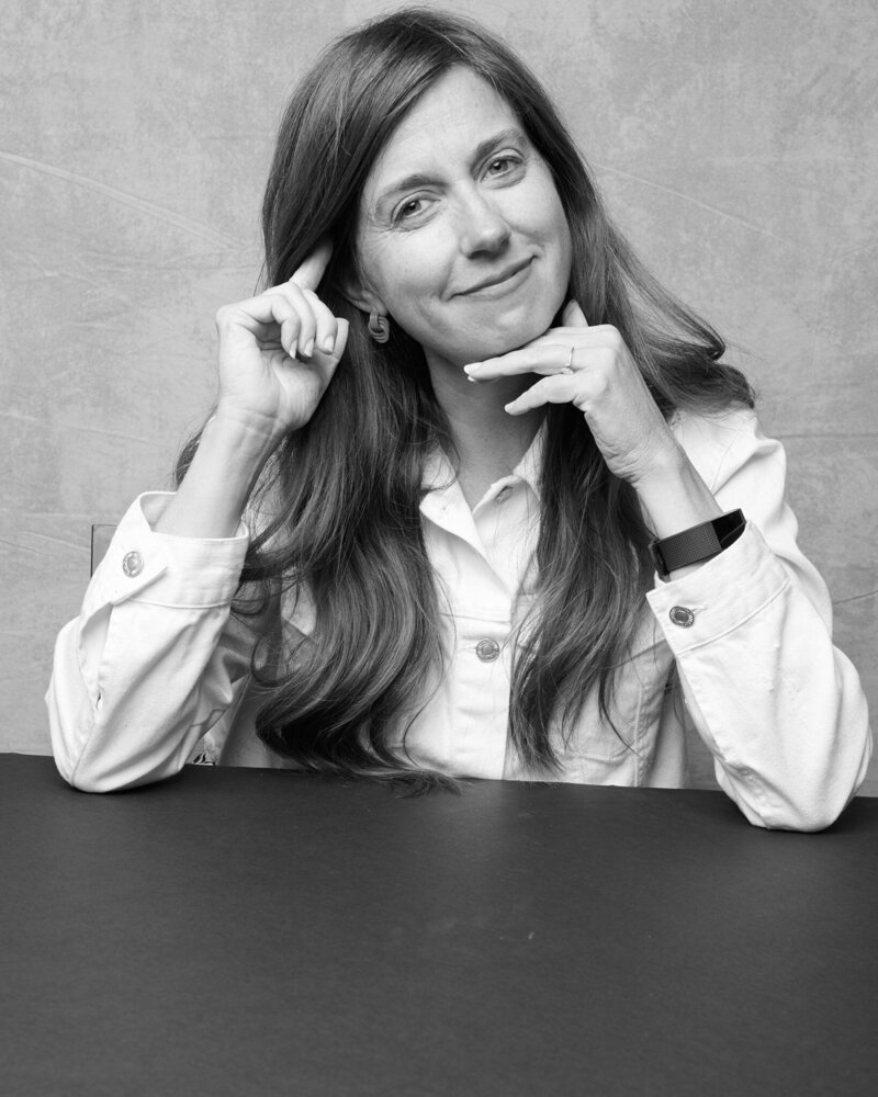 Black and white portrait of a woman in a white denim jacket, sitting at a table with a thoughtful, confident expression. She lightly touches her temple with one hand while resting the other near her chin, her long hair flowing naturally over her shoulders. A poised, unfiltered moment from 'THIS IS ME – A Photography Experience by Katie Campbell' in Atlanta, GA, celebrating authenticity and self-expression.