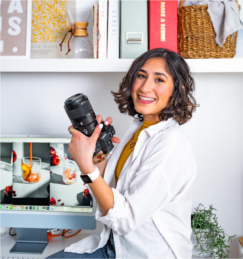 An image of Shahad sitting on her desk, holding her professional camera with a computer screen at the back showcasing her photography