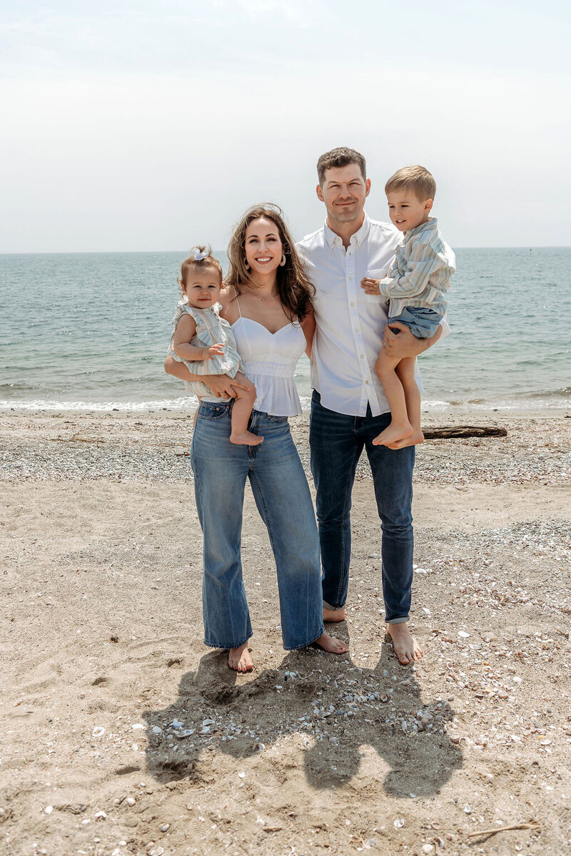 Happy family of four standing together on a sandy beach, with parents holding their young daughter and son against a backdrop of calm ocean waters and overcast sky