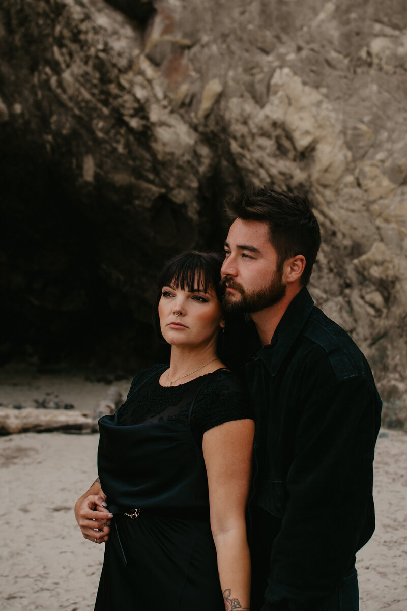 Cannon beach engagement session with the couple standing in front of a light rock formation in their black clothing. He is holding her from behind as they look into the distance.