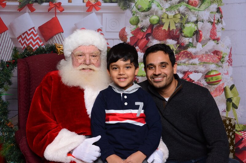 A heartwarming holiday photo of a child and parent sitting with Santa Claus in front of a beautifully decorated Christmas tree and festive backdrop