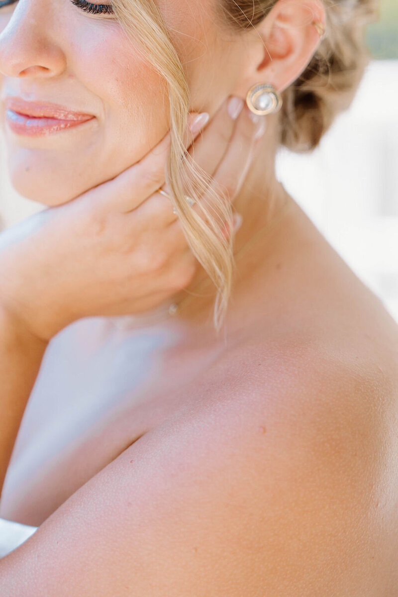 Newport Wedding Photographer | Close-up of a bride's face and shoulder, highlighting her delicate smile and elegant updo. She gently touches her cheek, showcasing a pearl earring.