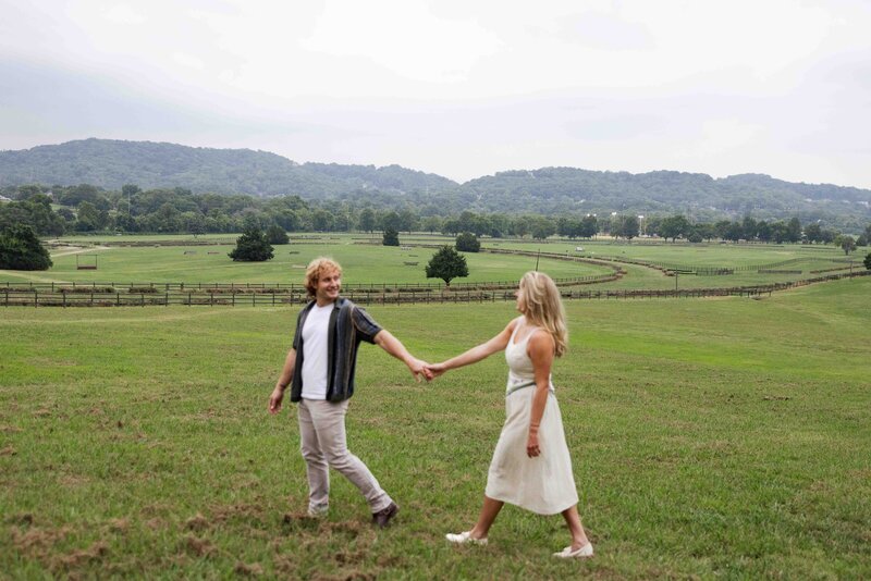 Man leads women in dress across green field while smiling back at her