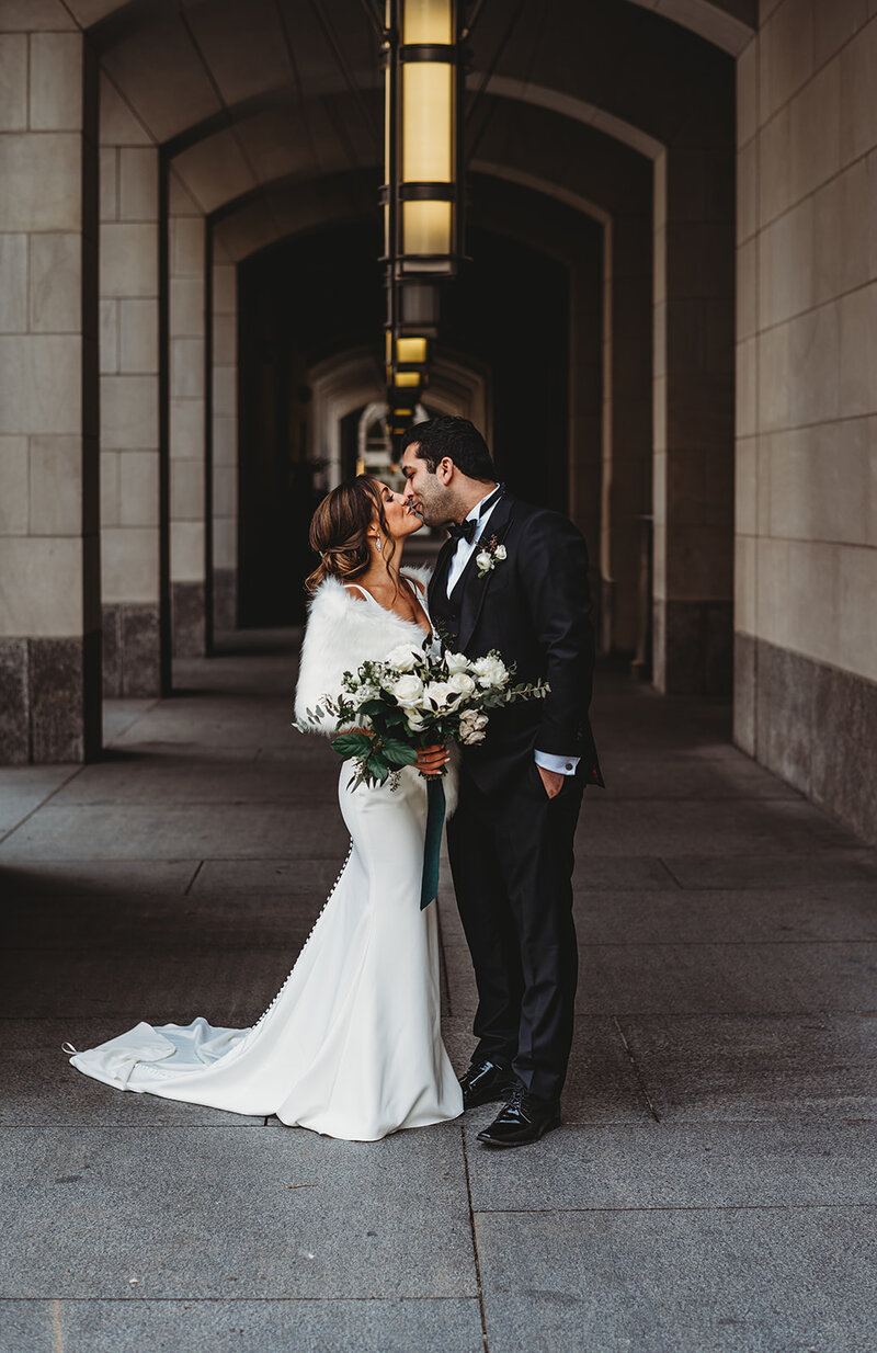 Winter wedding shot by Maryland wedding photographer with bride and groom kissing one another in a corridor of a stone building with bride and a satin gown wearing faux fur coat