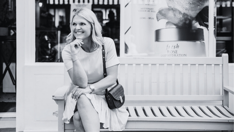 Emily Collins smiling in front of a brick wall, wearing a white tee and gold necklace.