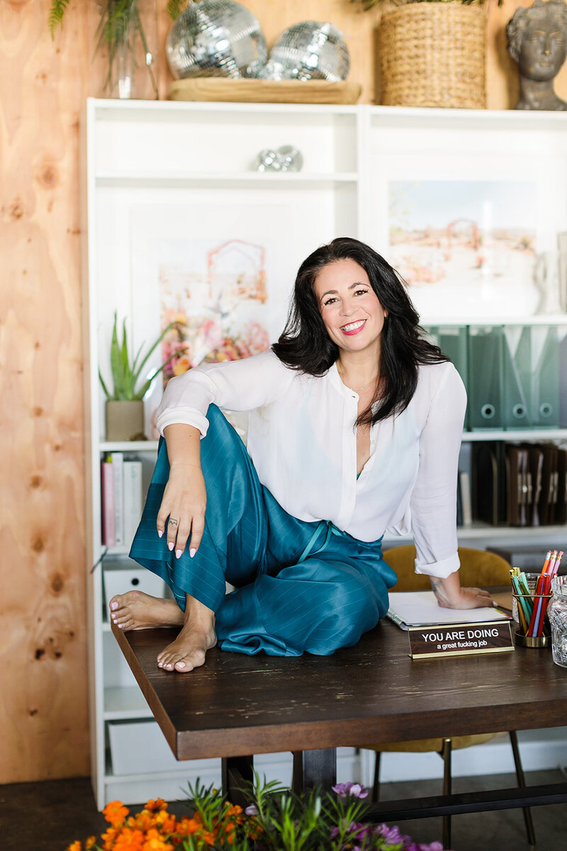 woman in silk blue pants and white flowy shirt sits cross legged on a desk and smiles