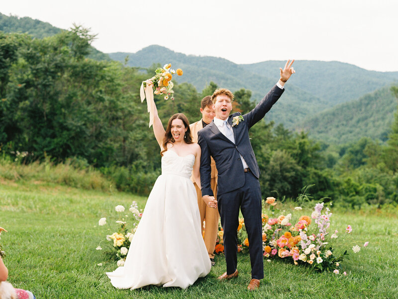 The bride and groom hold hands and raise their other hands in celebration of their wedding at the venue Paint Rock Farm by My Sun and Stars Co.