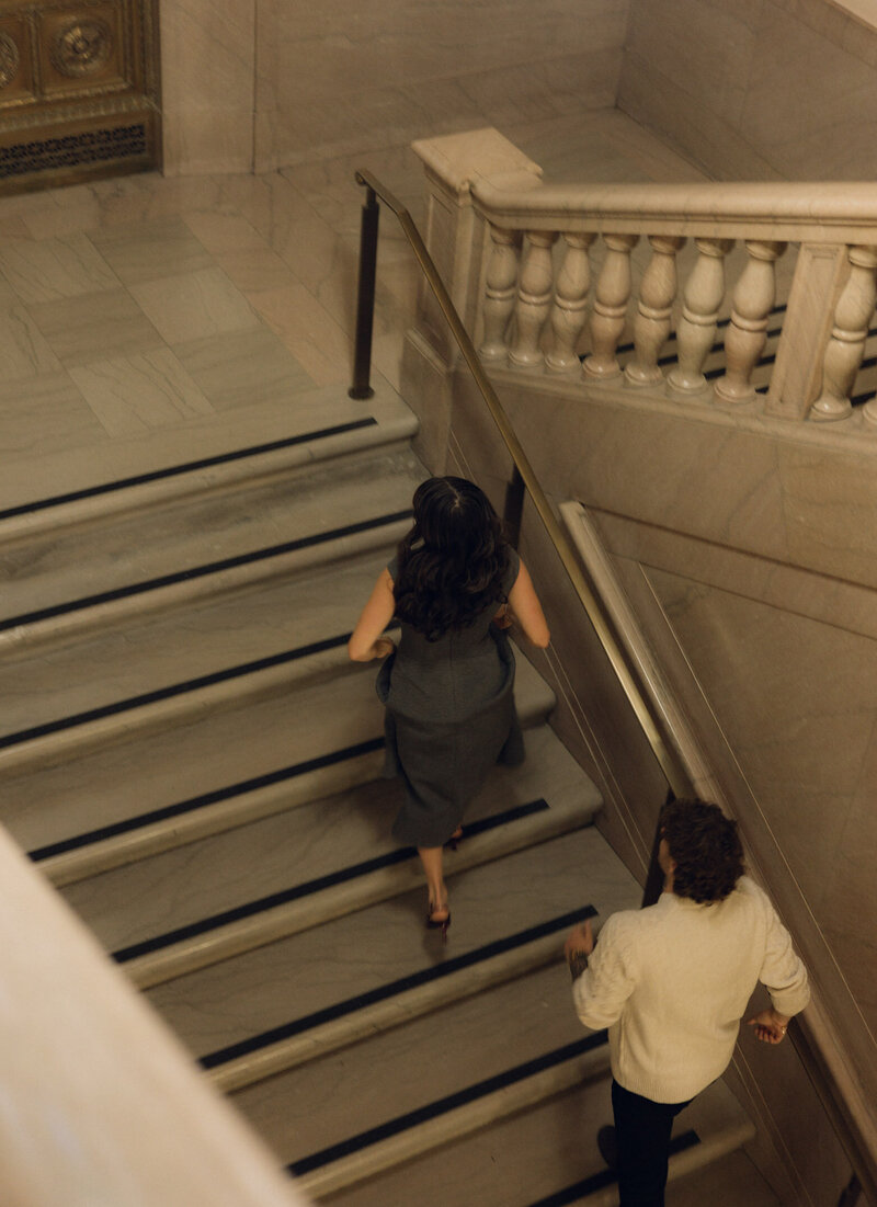 A couple runs up a staircase in a cinematic engagement photo at a library.