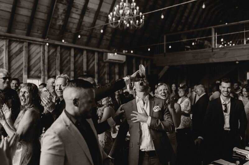 Wedding celebration with a guest pouring a shot into another guest’s mouth while others dance and clap around them.