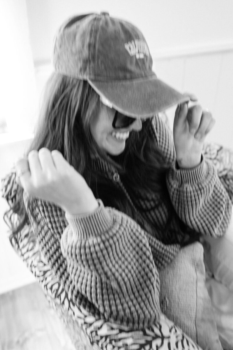 black and white shot of Annie tipping her baseball cap, wearing sunglasses and a sweater