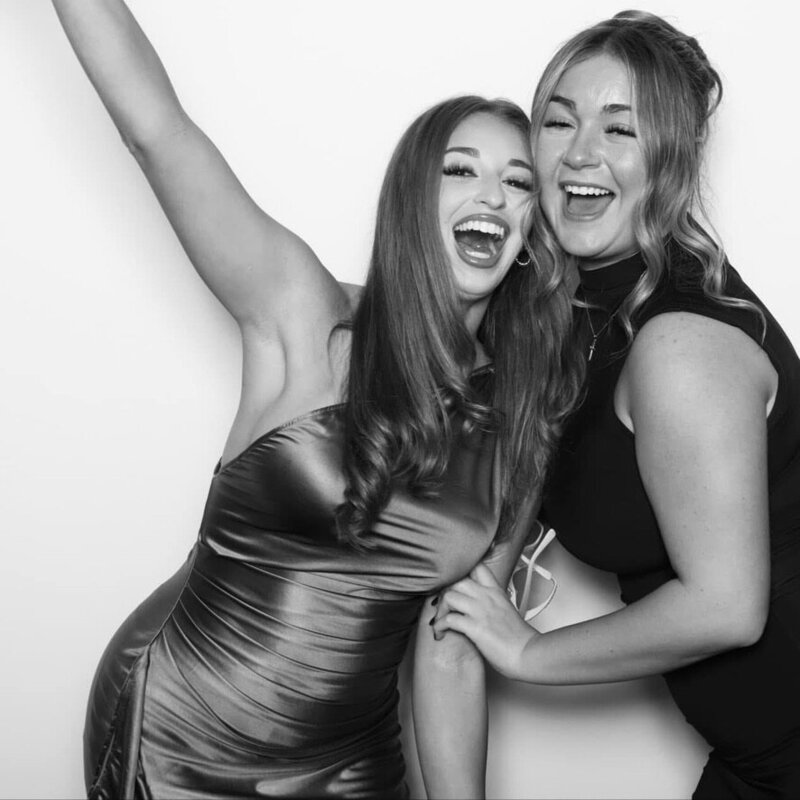 Two women laughing and embracing in front of a white backdrop. One raises her arm in excitement, and both appear joyful and energetic.
