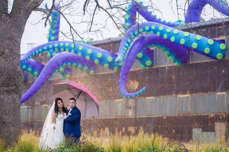 Couple standing in the rain in front of an octopus arm art installation at the Philadelphia Naval Yard, highlighting creative wedding style.
