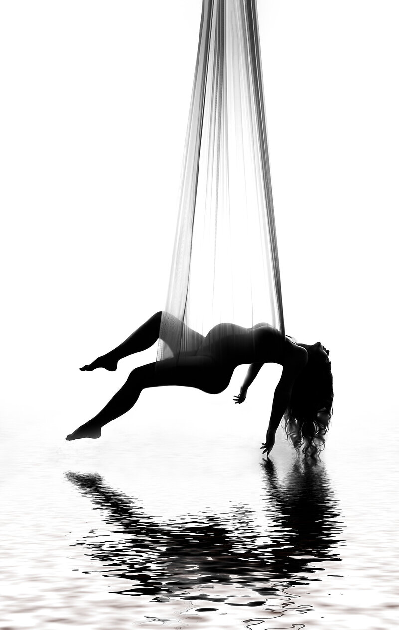 Premiere luxury studio maternity photography session at top Philadelphia, Pennsylvania studio - Mom is suspended from silks from the ceiling with her legs and arms stretched toward the ground. There is the illusion she is suspended over water with her reflection in the water.