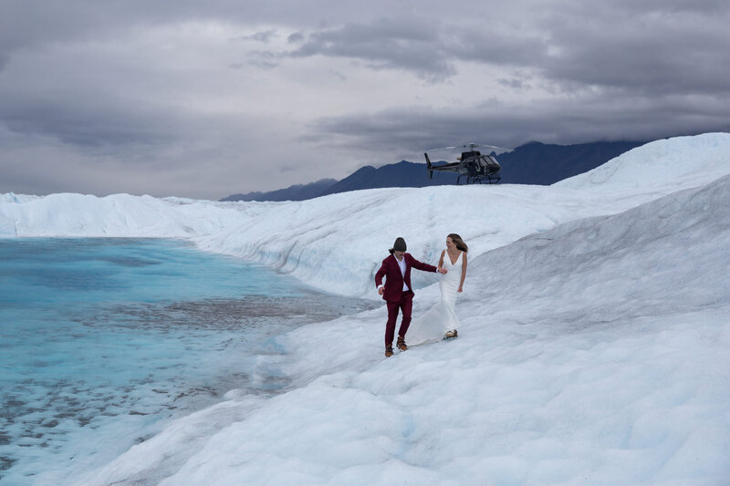 A bride wearing a white dress and a groom wearing a maroon suit and a beanie walk hand in hand on a glacier with a bright blue glacier pool next to them.