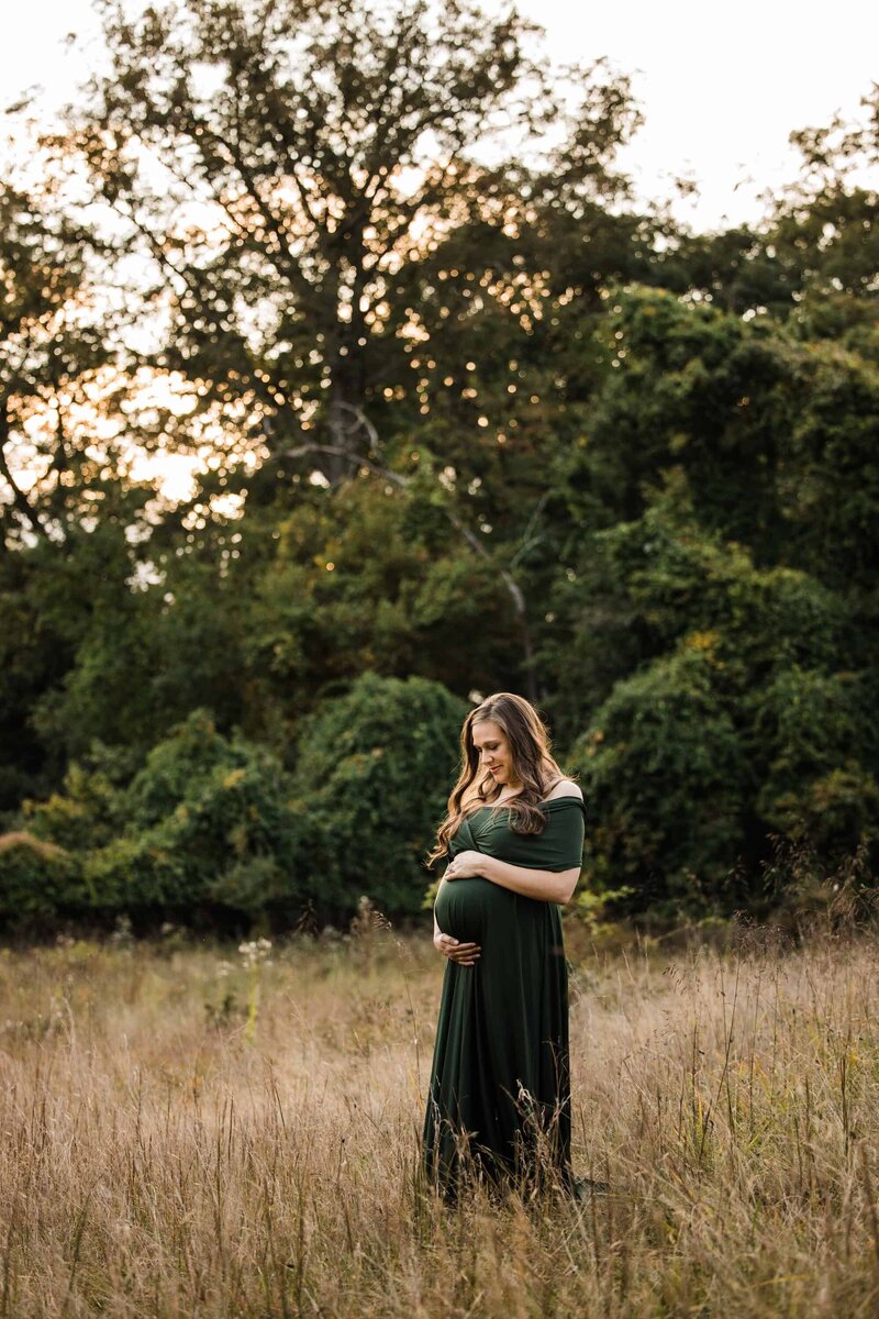 A pregnant woman in an olive green dress poses in front of a stone wall during a family photography session in Pittsburgh.