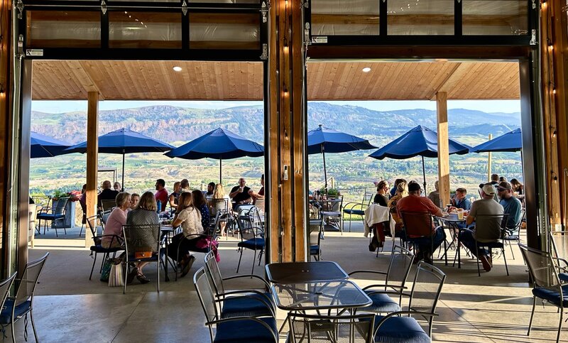 Standing inside of the tasting room at Union Hill Cider Co. Inside there are tables and chairs, then through the retractable walls there are people sitting on the patio eating and drinking. They are sitting under umbrellas looking out over the Wenatchee Valley. There are beautiful mountains in the background.