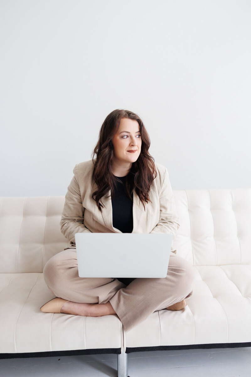 Woman sitting cross-legged on a modern white couch, working on a laptop – professional brand and web designer helping women entrepreneurs build bold brands