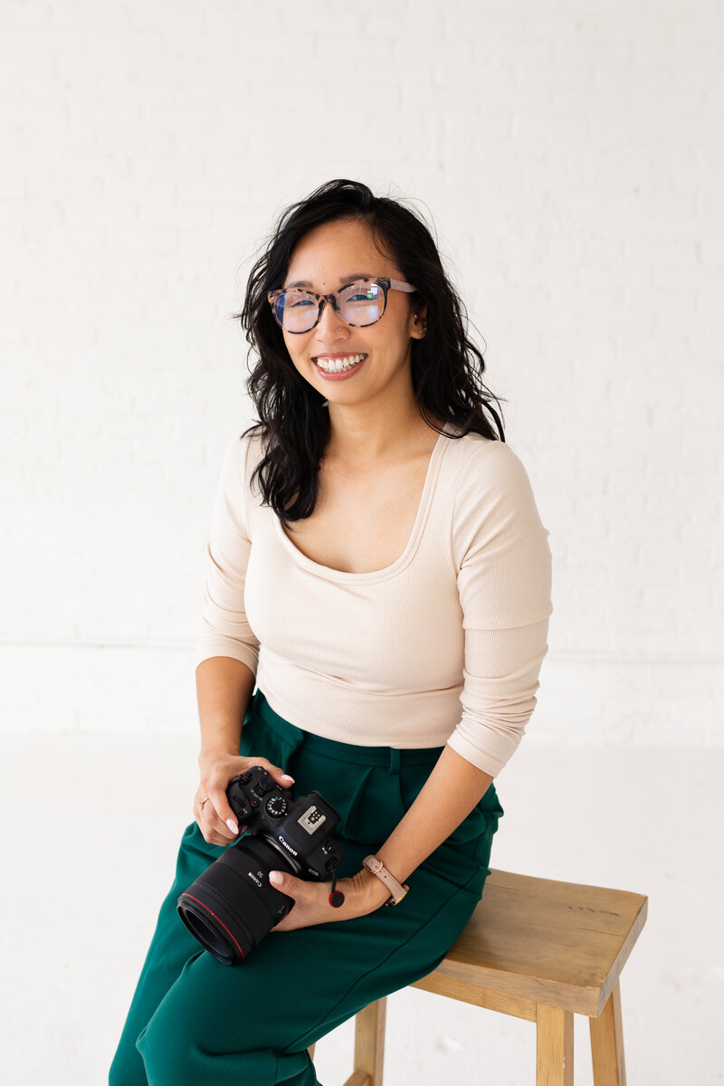 Anh smiling with a camera on a stool in a white top and teal pants